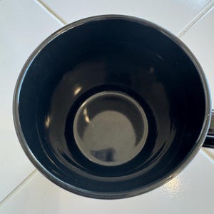 May include: A black ceramic mug, viewed from above, showcasing its interior. The mug has a glossy finish and a simple, classic design. The interior is a deep black, with a slightly lighter black circle at the bottom.