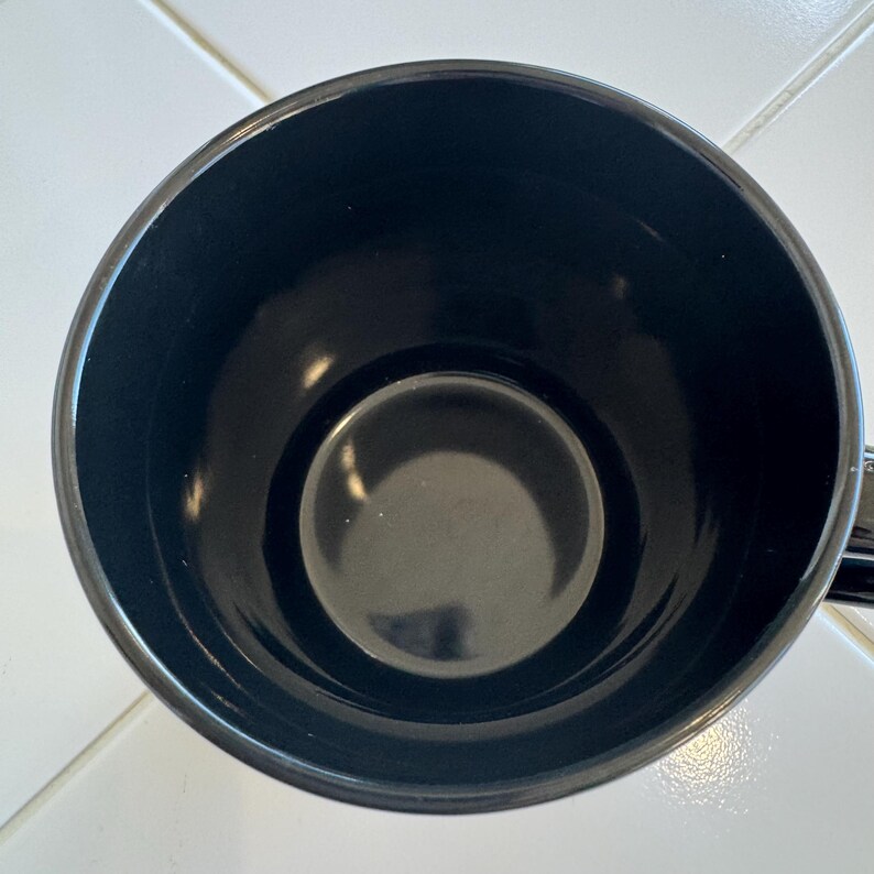 May include: A black ceramic mug, viewed from above, showcasing its interior. The mug has a glossy finish and a simple, classic design. The interior is a deep black, with a slightly lighter black circle at the bottom.
