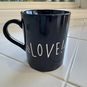 May include: A navy blue ceramic mug with a curved handle. The word "LOVE!" is written in white, stylized lettering on the side of the mug. The mug is sitting on a white tiled surface.