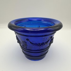 May include: A cobalt blue glass planter featuring a wide, rounded rim and a tapered body. The exterior surface is decorated with a raised relief pattern consisting of interconnected swags and circular motifs, complemented by horizontal ring details.