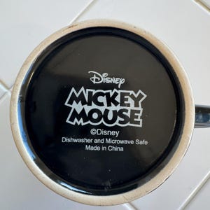May include: Black ceramic mug featuring the Disney Mickey Mouse logo in white. The mug's base includes the Disney logo, and text indicating it's dishwasher and microwave safe, made in China.