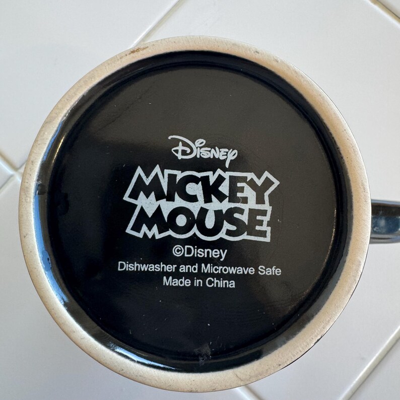 May include: Black ceramic mug featuring the Disney Mickey Mouse logo in white. The mug's base includes the Disney logo, and text indicating it's dishwasher and microwave safe, made in China.