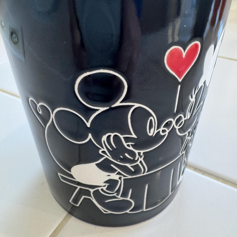 May include: A black tumbler featuring a white embossed design of Mickey Mouse holding a red heart-shaped balloon. The tumbler has a glossy finish and a cylindrical shape. The design includes the words "Love" and "Forever".