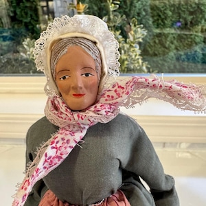 May include: A vintage doll with a painted face, wearing a white lace bonnet, a gray dress, and a pink floral scarf. The doll's face has painted features, including red lips and dark eyebrows. The doll's dress has a pink skirt.