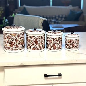 May include: A set of four cylindrical white enamel canisters of varying sizes, each featuring a detailed brown and gold paisley pattern. Every canister has a matching lid with a small black knob handle and a silver-toned rim at the top edge.