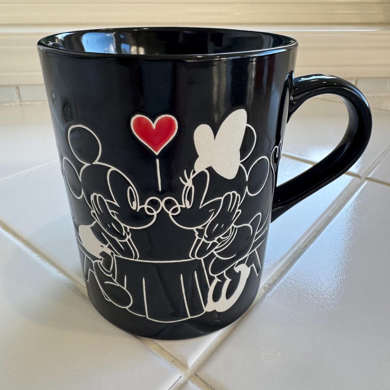 May include: Black ceramic mug featuring a white outline of Mickey and Minnie Mouse kissing, with a red heart between them. The mug has a curved handle and a glossy finish, perfect for coffee or tea.