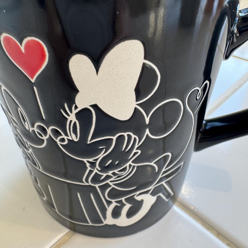 May include: Black ceramic mug featuring a white embossed design of Minnie Mouse. A red heart-shaped balloon is also part of the design. The mug has a black handle and is likely for hot beverages.