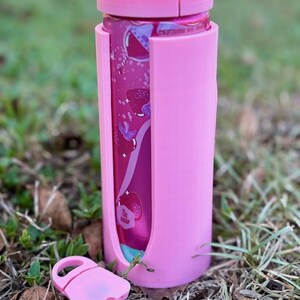 Puede incluir: Un enfriador de latas rosa con una tapa a juego y un cierre rosa separado. El enfriador est&aacute; dise&ntilde;ado para contener una lata, visible a trav&eacute;s de un recorte. La lata tiene un dise&ntilde;o rosa y morado. El enfriador est&aacute; sobre hierba.