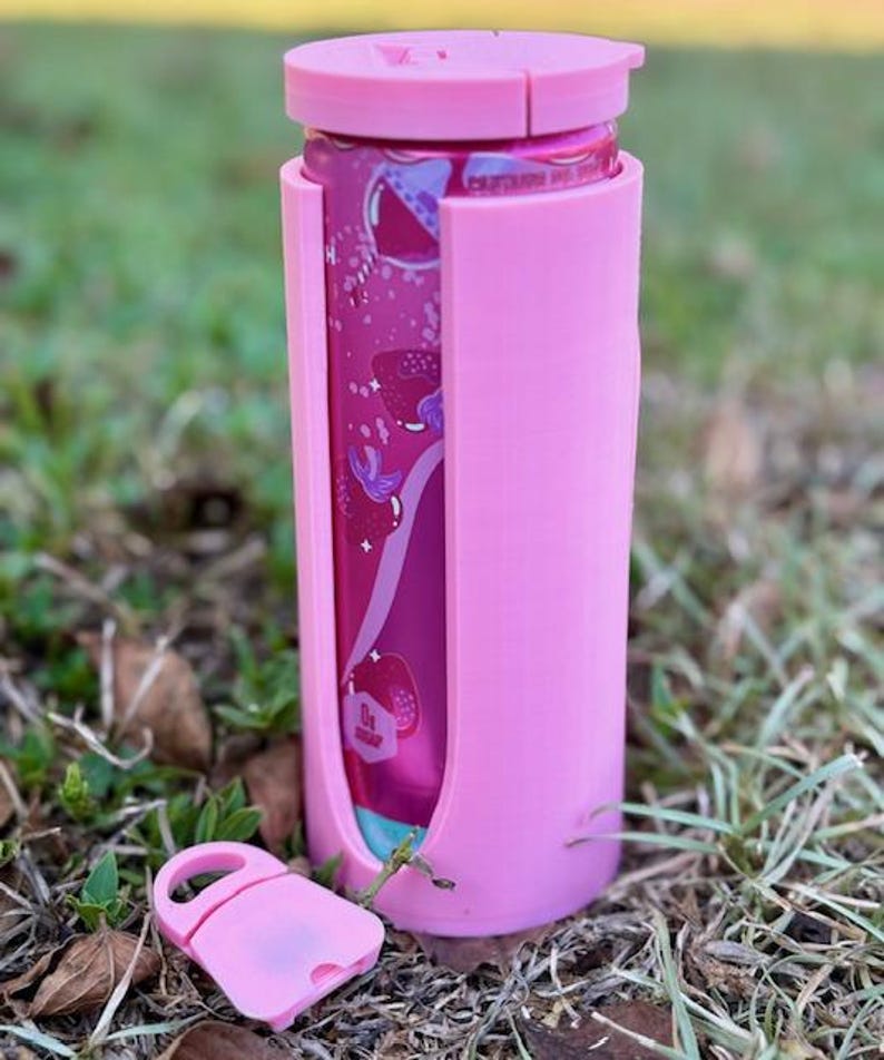 Puede incluir: Un enfriador de latas rosa con una tapa a juego y un cierre rosa separado. El enfriador est&aacute; dise&ntilde;ado para contener una lata, visible a trav&eacute;s de un recorte. La lata tiene un dise&ntilde;o rosa y morado. El enfriador est&aacute; sobre hierba.