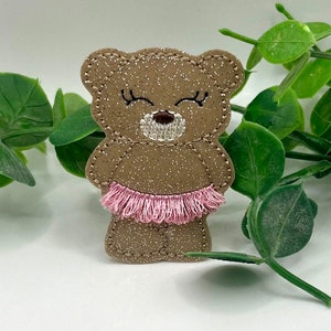 May include: A brown glitter bear wearing a pink tutu. The bear has closed eyes and a brown nose.