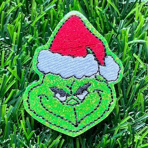 May include: Green Grinch character wearing a red and white Santa hat. The Grinch is made of green glitter fabric with black stitching.