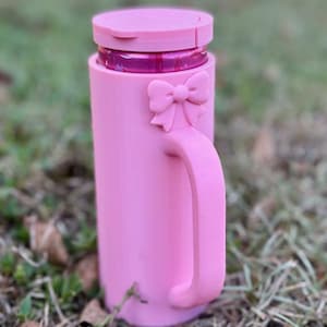 Puede incluir: Una taza rosa con asa y detalle de lazo. La taza tiene tapa y parece ser de pl&aacute;stico. La taza est&aacute; sobre hierba.