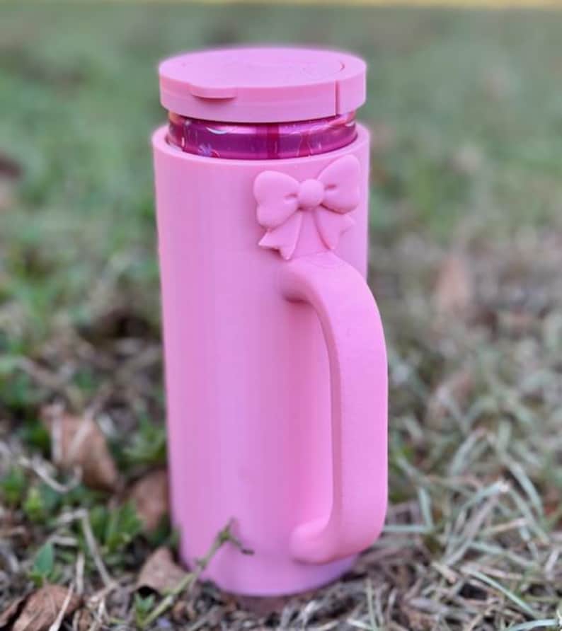 Puede incluir: Una taza rosa con asa y detalle de lazo. La taza tiene tapa y parece ser de pl&aacute;stico. La taza est&aacute; sobre hierba.