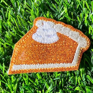 May include: A brown and orange glitter pumpkin pie patch with a white whipped cream topping. The patch is sewn with white thread and has a white crust.