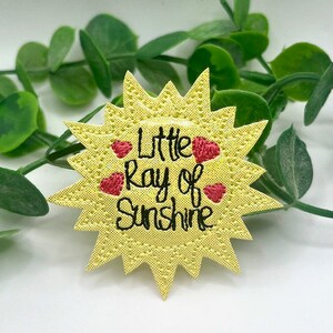 May include: A yellow sun-shaped patch with the text "Little Ray of Sunshine" in black script, and three red heart accents. The patch has a textured surface and is surrounded by green leaves.