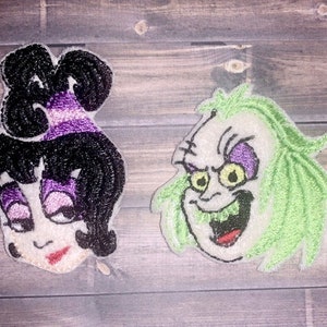 May include: Two embroidered iron-on patches featuring cartoon characters. The first patch is a purple-haired woman with a white face and black eyes. The second patch is a green-haired man with a white face and purple eyes.