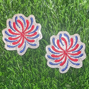 May include: Two red, white, and blue sparkly firework-shaped iron-on patches. The patches are on a green grass background.