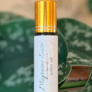 Puede incluir: Un frasco de aceite en roll-on con tapa dorada y etiqueta blanca. La etiqueta dice "Migraine Ease roller oil" en letra cursiva con un diseño de flecha decorativa. El frasco está sobre un fondo borroso de hojas verdes.
