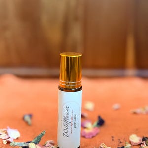 May include: A perfume bottle with a gold cap and a white label reading "Wildflower perfume." The bottle is set on an orange surface, surrounded by dried flowers. The background is a blurred wooden texture.