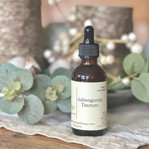 May include: A brown glass bottle of Ashwaganda Tincture with a black dropper. The label is cream-colored with black text. The bottle is surrounded by green and white decorative foliage on a neutral surface.