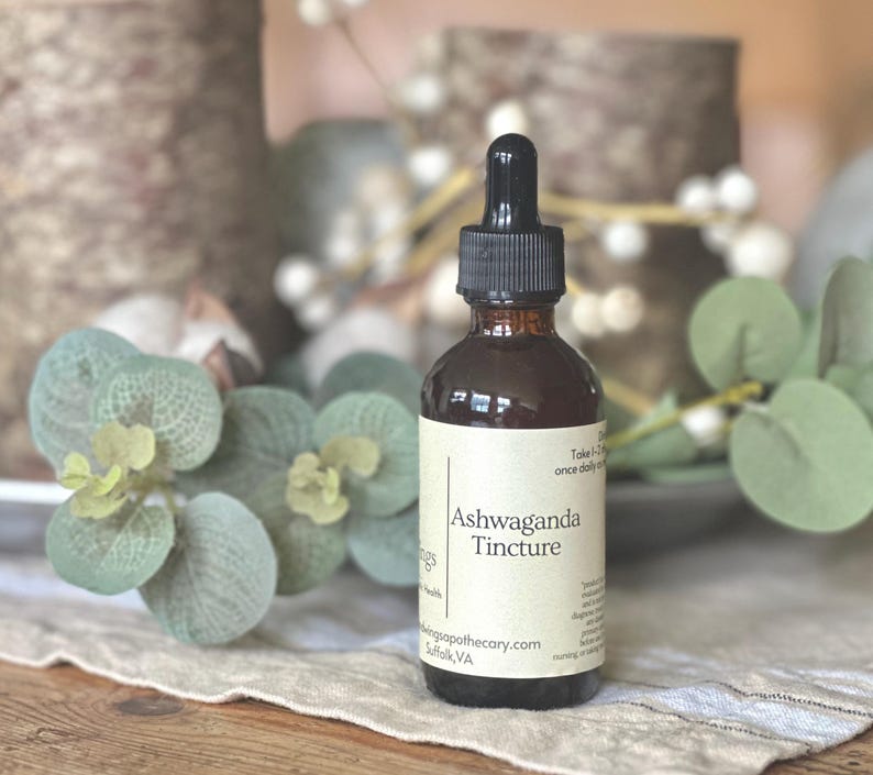 May include: A brown glass bottle of Ashwaganda Tincture with a black dropper. The label is cream-colored with black text. The bottle is surrounded by green and white decorative foliage on a neutral surface.