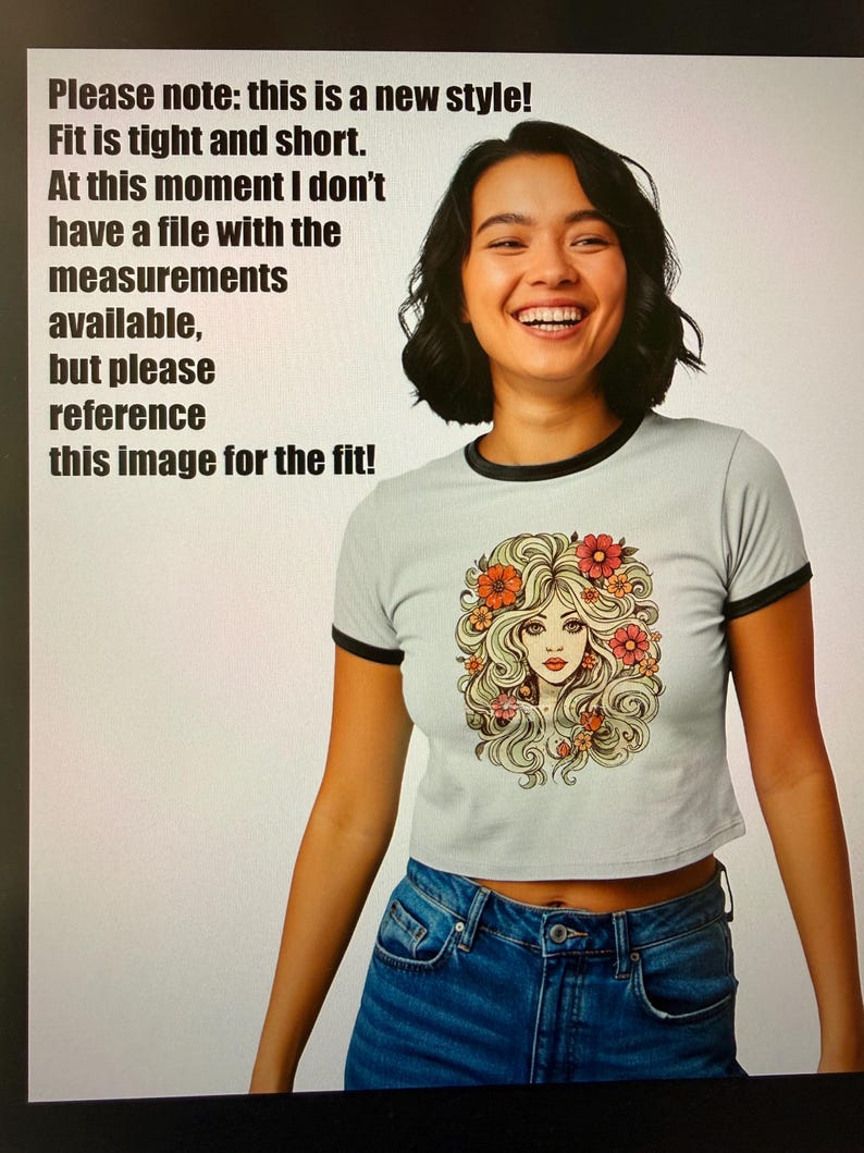 May include: A light gray crop top with black trim and a graphic of a woman with flowers in her hair. The text on the image says, "Please note: this is a new style! Fit is tight and short. At this moment I don't have a file with the measurements available, but please reference this image for the fit!"