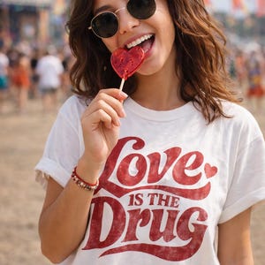 Könnte beinhalten: Eine Person mit Sonnenbrille und einem weißen T-Shirt mit dem roten Text "Love is the Drug" lächelt und hält einen herzförmigen Lolli. Die Person trägt ein rotes Armband. Der Hintergrund ist verschwommen mit Menschen und bunten Fahnen.