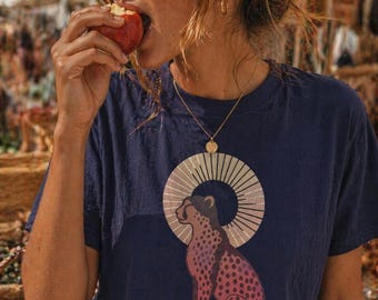 May include: Navy blue t-shirt featuring a cheetah design with a halo. The cheetah is depicted in shades of pink and purple with black spots. The shirt has a round neck and short sleeves. A gold necklace is visible.