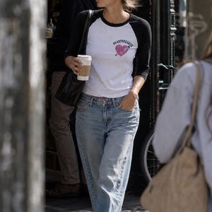May include: A white and black raglan sleeve t-shirt featuring a pink heart graphic with a dagger through it and the text HEARTBREAKER printed above. The shirt is paired with light blue denim jeans and grey athletic sneakers.