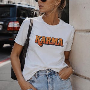 May include: White t-shirt with the word "KARMA" in orange and yellow block letters. The shirt is paired with light blue denim shorts. The person is wearing black sunglasses and has their hair up in a bun.