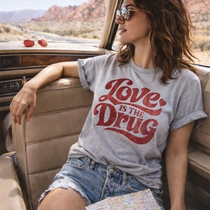 May include: A gray t-shirt with the text "Love is the Drug" in red script. The person is wearing sunglasses and denim shorts, holding a map, and sitting in a car. The background shows a desert landscape.