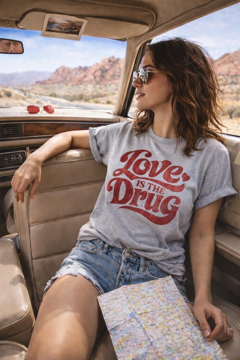 May include: A gray t-shirt with the text "Love is the Drug" in red script. The person is wearing sunglasses and denim shorts, holding a map, and sitting in a car. The background shows a desert landscape.