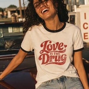 Puede incluir: Una camiseta blanca tipo ringer con ribetes negros en el cuello y las mangas. La parte delantera presenta letras rojas con efecto desgastado que dicen Love is the Drug, con un pequeño icono de corazón integrado en el diseño.