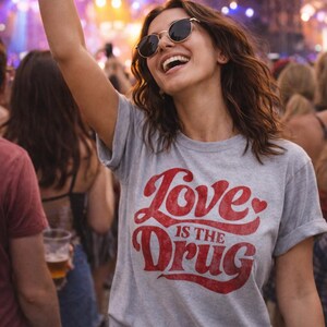 May include: A gray t-shirt with the text "Love is the Drug" in red script. The person is wearing sunglasses and smiling with their arm raised in a crowd at a concert.