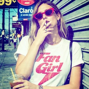 May include: A person wearing red cat-eye sunglasses and a white t-shirt with the text "Fan Girl" in red. The person is smoking a cigarette and holding a beverage. The background includes street signs and a metal shutter.