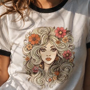 May include: White t-shirt with a black trim around the collar and sleeves. The shirt features a vintage-style illustration of a woman with long, flowing blonde hair adorned with colorful flowers in shades of orange and pink.