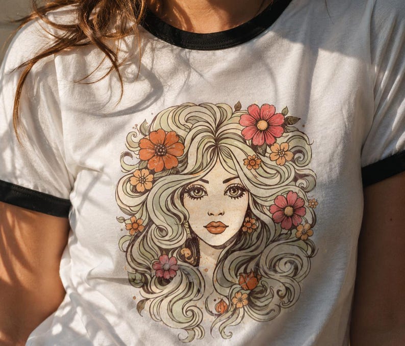 May include: White t-shirt with a black trim around the collar and sleeves. The shirt features a vintage-style illustration of a woman with long, flowing blonde hair adorned with colorful flowers in shades of orange and pink.