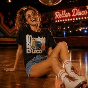 Może przedstawiać: Osoba w czarnym t-shircie z grafiką "Moonlight Roller Disco", dżinsowych szortach i rolkach. Rolki są białe z czerwonymi sznurowadłami i pomarańczowymi k&oacute;łkami. W tle kula dyskotekowa i neonowy znak "Roller Disco".
