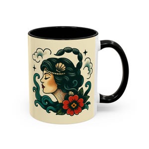 May include: A white ceramic mug with a black interior and handle. The mug features a vintage-style illustration of a woman with dark hair, a scorpion, and a red flower. The design is set against a cream background.