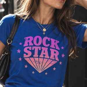 May include: A royal blue short-sleeved t-shirt featuring the text ROCK STAR in a distressed pink font. Below the text is a pink and orange sunburst graphic, surrounded by small pink stars arranged in a circular pattern on the front of the shirt.
