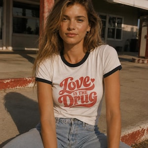 Könnte beinhalten: Weißes Ringer-T-Shirt mit schwarzem Besatz und dem Text "Love is the Drug" in roter Schrift. Das Model trägt eine hellblaue Jeans. Das Bild ist im Freien bei natürlichem Licht aufgenommen.