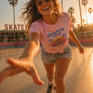 May include: A woman wearing a pink t-shirt with "Rollerskating Rink Los Angeles" text, denim shorts, and roller skates. She is smiling and wearing sunglasses. Palm trees and a "SKATE" sign are in the background.