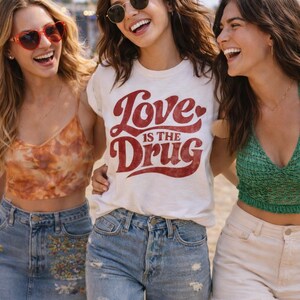 May include: Three smiling people wearing casual clothing. One person wears a white t-shirt with the text "Love is the Drug" in red. Others wear sunglasses, a tie-dye top, and a green top. They are standing outside.