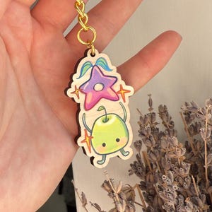 May include: A colorful keychain featuring a star and a green apple-like character with a gold chain and ring. The star is purple and pink, and the apple character has a smiling face. The keychain is a fun accessory.
