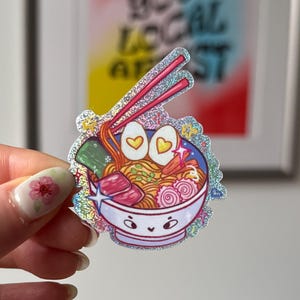 Puede incluir: Una pegatina holográfica con una ilustración de un cuenco de ramen de dibujos animados con palillos. El cuenco tiene una cara sonriente y contiene fideos, huevos y otros ingredientes. El texto "SUPPORT YOUR LOCAL ARTIST" es visible en el fondo.