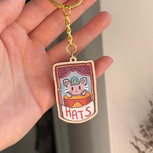 May include: A wooden keychain with a cartoon mouse wearing a hat, set within a stage-like frame. The word "HATS" is below. The keychain has a gold-coloured ring and chain.