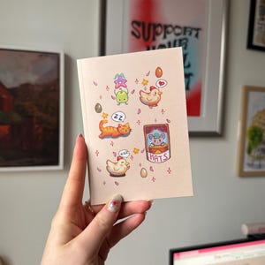Op de afbeelding: Een roze wenskaart met cartoon illustraties van een kat, kippen, eieren en een kikker. De kaart heeft het woord "HATS" erop. De kaart wordt voor een muur met ingelijste kunstwerken gehouden.
