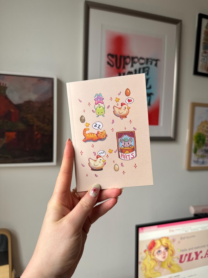 Op de afbeelding: Een roze wenskaart met cartoon illustraties van een kat, kippen, eieren en een kikker. De kaart heeft het woord "HATS" erop. De kaart wordt voor een muur met ingelijste kunstwerken gehouden.