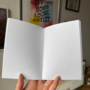 Op de afbeelding: Een open notitieboek met pagina's met stippenraster. Het notitieboek wordt opengehouden en toont de binnenpagina's. De achtergrond bevat een ingelijst kunstwerk met de tekst "SUPPORT YOUR LOCAL ARTIST".