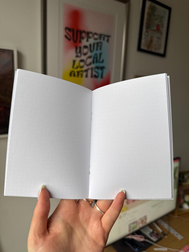 Op de afbeelding: Een open notitieboek met pagina's met stippenraster. Het notitieboek wordt opengehouden en toont de binnenpagina's. De achtergrond bevat een ingelijst kunstwerk met de tekst "SUPPORT YOUR LOCAL ARTIST".